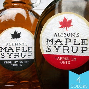 May include: Two glass bottles of maple syrup with labels. The labels are round and have a wood grain design. The labels are black and white with red accents. The text on the labels reads "Johnny's Maple Syrup" and "Alison's Maple Syrup".