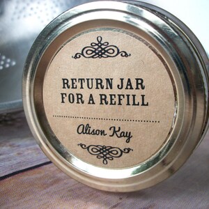 May include: A brown paper label on a silver jar lid with the text "RETURN JAR FOR A REFILL" and "Alison Kay" in black text.