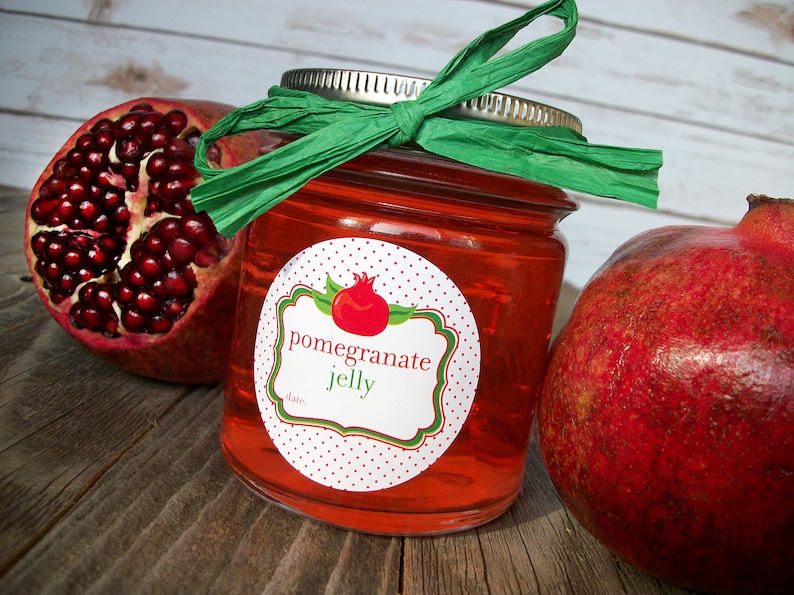May include: A jar of red pomegranate jelly with a white label that says "pomegranate jelly" and has a red pomegranate illustration. The jar is tied with a green ribbon and there are two whole pomegranates next to it.