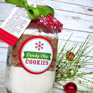 A mason jar filled with layered ingredients for Candy Chip Cookies. The jar is adorned with a red and white snowflake-patterned fabric lid cover, a green ribbon, and a tag with instructions. A round label reads "Candy Chip COOKIES".