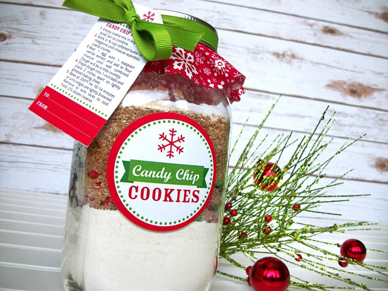 A mason jar filled with layered ingredients for Candy Chip Cookies. The jar is adorned with a red and white snowflake-patterned fabric lid cover, a green ribbon, and a tag with instructions. A round label reads "Candy Chip COOKIES".