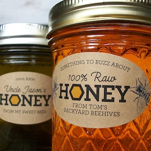May include: Two glass jars filled with amber-colored honey, each featuring a metallic lid and an oval-shaped tan label. The labels display the text "100% Raw HONEY" with bee illustrations and specific names like Uncle Jason's and Tom's.