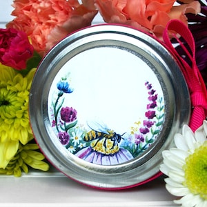 May include: A round, white label with a painted bee and flowers around the edge. The label is attached to a silver jar lid with a pink ribbon.