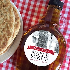 May include: A glass bottle of Tom's Pure Maple Syrup with a black cap and a handle. The label features a black and white illustration of trees and a banner with the text "Maple Syrup". Beside the bottle is a plate of pancakes on a red and white checkered tablecloth.