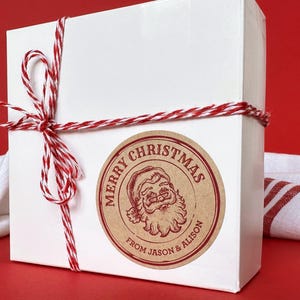 Puede incluir: Una caja de regalo cuadrada blanca atada con cordel de panadero rojo y blanco, con una pegatina redonda de papel kraft que muestra una ilustración roja de Santa Claus y el texto Merry Christmas from Jason & Alison en tinta roja.