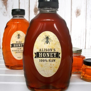 Vintage looking oval honey labels on the front of honey bottles filled with honey. Label art has a realistic honey bee up top with printed text for 100% Raw honey.