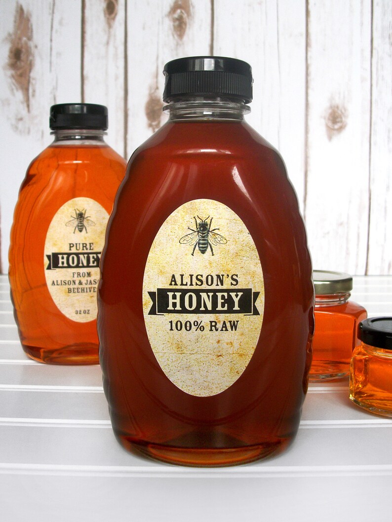 Vintage looking oval honey labels on the front of honey bottles filled with honey. Label art has a realistic honey bee up top with printed text for 100% Raw honey.
