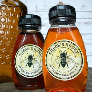 May include: Two glass bottles of honey with labels. The label on the left bottle says "Alison's Honey" and "From my backyard bees". The label on the right bottle says "Chuck's Honey" and "I got stung in the head for this!"