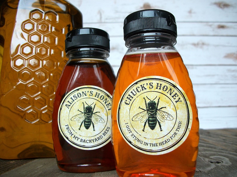 May include: Two glass bottles of honey with labels. The label on the left bottle says "Alison's Honey" and "From my backyard bees". The label on the right bottle says "Chuck's Honey" and "I got stung in the head for this!"