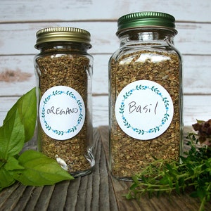 May include: Two glass jars with gold and green lids filled with dried herbs. The jars have white round labels with blue floral borders. The label on the left jar says "Oregano" and the label on the right jar says "Basil".