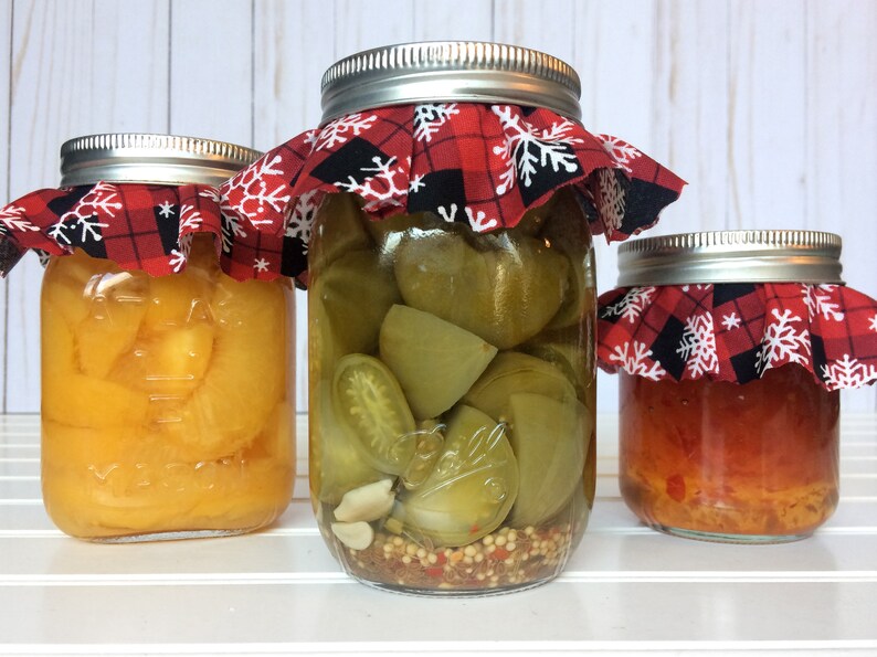 Puede incluir: Tres tarros de vidrio con tapas de tela a cuadros rojos y negros con copos de nieve blancos. Los tarros contienen pimientos amarillos, tomates verdes y pimientos rojos en conserva.