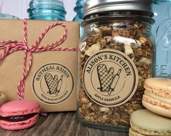 May include: A glass jar filled with apple granola and a brown box tied with red and white twine. Both feature circular labels with the text Alison's Kitchen, an oven mitt, and a rolling pin. A pink macaron and two beige macarons are placed nearby.