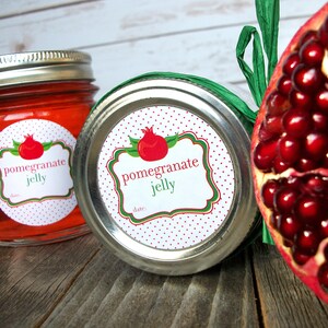 May include: Two glass jars with red pomegranate jelly. The jars have white round labels with red polka dots and green accents. The labels say "pomegranate jelly" and have a space for a date. A red pomegranate fruit is visible in the background.