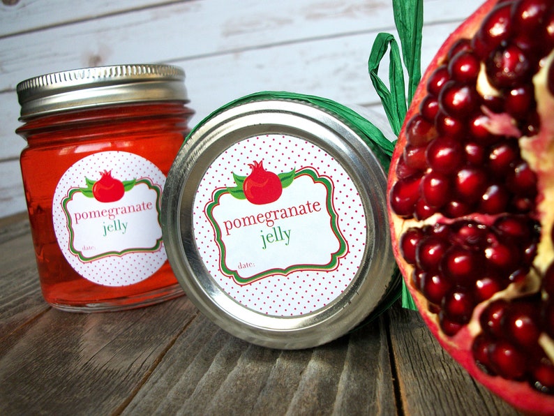 May include: Two glass jars with red pomegranate jelly. The jars have white round labels with red polka dots and green accents. The labels say "pomegranate jelly" and have a space for a date. A red pomegranate fruit is visible in the background.