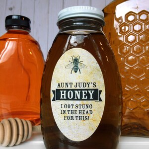 Vintage looking oval honey label on the front of honey bottle filled with honey. Label art has a realistic honey bee up top with printed text for honey. In the background are jars filled with honey and wooden honey dippers.