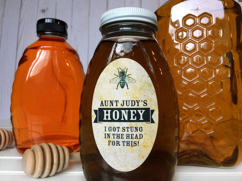 Vintage looking oval honey label on the front of honey bottle filled with honey. Label art has a realistic honey bee up top with printed text for honey. In the background are jars filled with honey and wooden honey dippers.