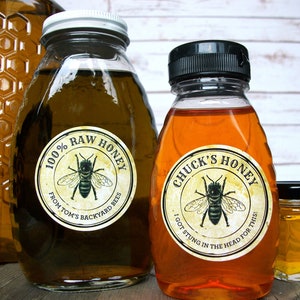 May include: Three glass jars of honey with labels. The largest jar is clear and has a label that reads "100% Raw Honey from Tom's Backyard Bees". The middle jar is amber colored and has a label that reads "Chuck's Honey I got stung in the head for this!". The smallest jar is a clear square jar with a black lid.
