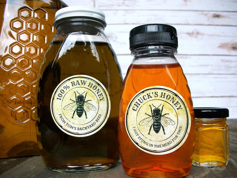 May include: Three glass jars of honey with labels. The largest jar is clear and has a label that reads "100% Raw Honey from Tom's Backyard Bees". The middle jar is amber colored and has a label that reads "Chuck's Honey I got stung in the head for this!". The smallest jar is a clear square jar with a black lid.