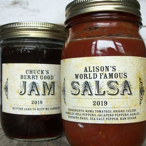 May include: Two glass jars with labels. The jar on the left is filled with dark brown jam and has a label that reads "Chuck's Berry Good Jam 2019 Return Jars to Keep Me Jammin". The jar on the right is filled with red salsa and has a label that reads "Alison's World Famous Salsa 2019 Ingredients: Roma Tomatoes, Onions, Celery, Parsley, Bell Peppers, Jalapeno Peppers, Garlic, Oregano, Basil, Sea Salt, Pepper, Raw Sugar".