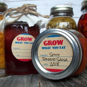 May include: Four glass jars with metal lids and labels. The labels are brown with white text and a red and blue border. The text on the labels reads "Grow What You Eat". One jar has a label that says "Spicy Tomato Salsa" and "2018".
