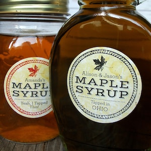 May include: Two glass jars of maple syrup with labels. The label on the left jar says "Amanda's Maple Syrup" and "Yeah, I Tapped This!" The label on the right jar says "Alison & Jason's Maple Syrup" and "Tapped in OHIO".
