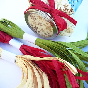 May include: Three spools of paper raffia ribbon in green, red, and cream colors. The ribbon is wrapped around a white cardboard core.