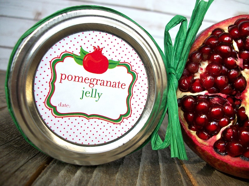 May include: A white jar with a red and green polka dot label that says "pomegranate jelly" and "date:". The jar is next to a half of a pomegranate.
