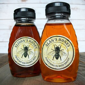 May include: Two glass honey bottles with black lids and labels. The labels are round and feature a bee illustration. The left bottle's label reads "Alison's Honey From My Backyard Bees". The right bottle's label reads "Alan's Honey Something to Buzz About!"