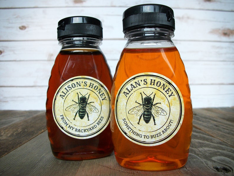 May include: Two glass honey bottles with black lids and labels. The labels are round and feature a bee illustration. The left bottle's label reads "Alison's Honey From My Backyard Bees". The right bottle's label reads "Alan's Honey Something to Buzz About!"