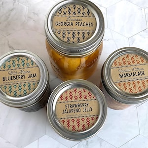 Puede incluir: Cuatro frascos de vidrio con tapas metálicas que contienen productos alimenticios. Las etiquetas dicen Bourbon Georgia Peaches, Wild Maine Blueberry Jam, Vanilla Citrus Marmalade y Strawberry Jalapeno Jelly, cada una con un patrón de árboles.
