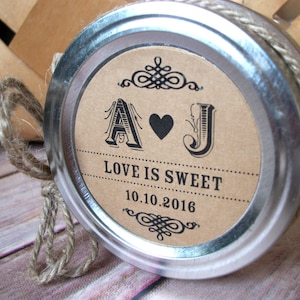 May include: A mason jar lid with a decorative label. The label is tan with black text and features the letters "A" and "J" with a heart symbol, and the words "LOVE IS SWEET" and the date "10.10.2016". The lid is wrapped with twine.