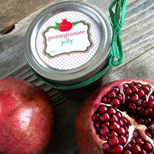 May include: A glass jar with a red and white polka dot label that says "pomegranate jelly" with a green border. The jar is tied with a green ribbon. There are two pomegranates next to the jar, one whole and one cut open.