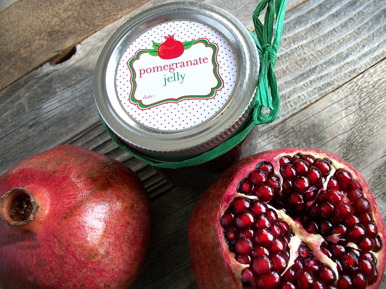 May include: A glass jar with a red and white polka dot label that says "pomegranate jelly" with a green border. The jar is tied with a green ribbon. There are two pomegranates next to the jar, one whole and one cut open.