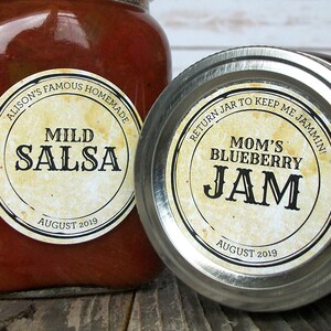 May include: Two glass jars of homemade preserves. One jar contains Mild Salsa, the other Mom's Blueberry Jam. Both jars have labels with text and the date August 2019. The salsa jar is open, while the jam jar is sealed.
