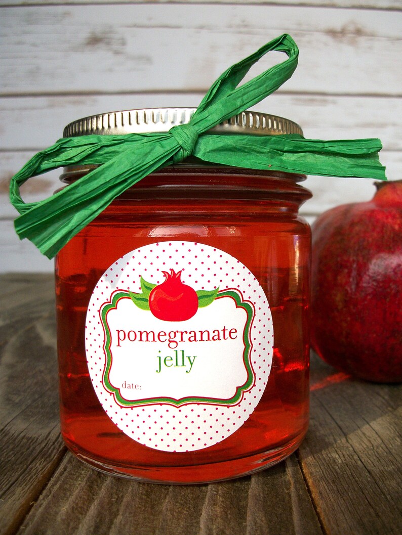 May include: A glass jar filled with red pomegranate jelly, topped with a green ribbon bow. The jar has a white label with red polka dots and the text "pomegranate jelly" and "date:".