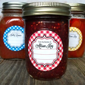 May include: Three glass jars with red, blue, and yellow gingham labels. The labels have a white border and say "From the Kitchen of" followed by a name. The jars are filled with red, orange, and a dark red substance.