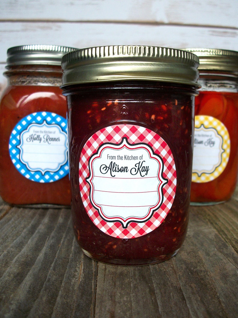 May include: Three glass jars with red, blue, and yellow gingham labels. The labels have a white border and say "From the Kitchen of" followed by a name. The jars are filled with red, orange, and a dark red substance.