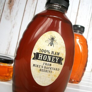 Vintage looking oval honey label on the front of honey bottle filled with honey. Label art has a realistic honey bee up top with printed text for 100% Raw honey. In the background are jars filled with honey.