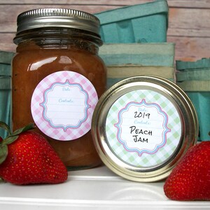 May include: Two glass jars with lids, filled with a brown substance. The jars have round, pink and blue gingham labels with white text that reads "Date: Contents". One label has the date "2019" and the words "Peach Jam".