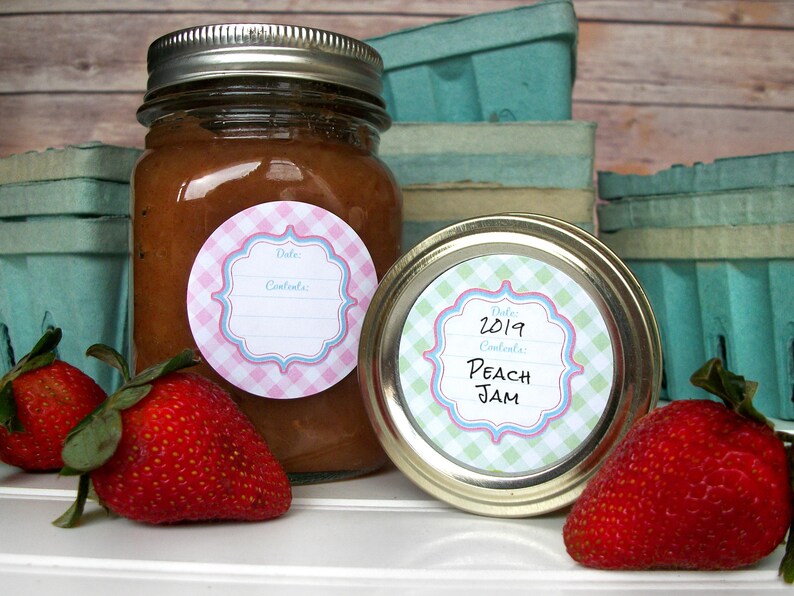 May include: Two glass jars with lids, filled with a brown substance. The jars have round, pink and blue gingham labels with white text that reads "Date: Contents". One label has the date "2019" and the words "Peach Jam".