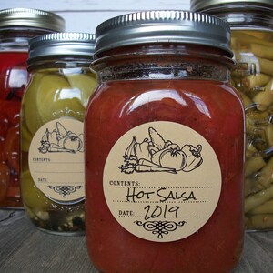May include: Three glass jars with silver lids and brown paper labels. The jars contain red salsa, green beans, and red peppers. The labels have the words "Contents: Hot Salsa Date: 2019" printed on them.