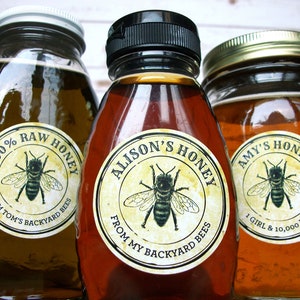 May include: Three glass jars of honey with labels. The labels are round and have a yellow background with black text. The text on the labels reads "100% RAW HONEY FROM TOM'S BACKYARD BEES", "ALISON'S HONEY FROM MY BACKYARD BEES", and "AMY'S HONEY 1 GIRL & 10,000 BEES". Each label also features a black and white illustration of a bee.