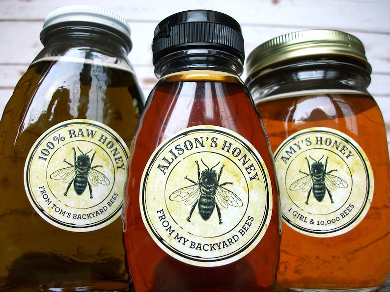 May include: Three glass jars of honey with labels. The labels are round and have a yellow background with black text. The text on the labels reads "100% RAW HONEY FROM TOM'S BACKYARD BEES", "ALISON'S HONEY FROM MY BACKYARD BEES", and "AMY'S HONEY 1 GIRL & 10,000 BEES". Each label also features a black and white illustration of a bee.