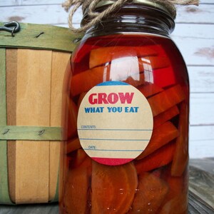 May include: A glass jar filled with orange slices in a red liquid. The jar has a brown paper label with blue and red trim that reads "GROW WHAT YOU EAT". The label also has blank spaces for "CONTENTS" and "DATE".