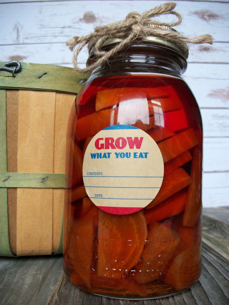 May include: A glass jar filled with orange slices in a red liquid. The jar has a brown paper label with blue and red trim that reads "GROW WHAT YOU EAT". The label also has blank spaces for "CONTENTS" and "DATE".