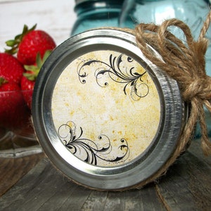 May include: A silver jar lid with a cream-colored label featuring a black floral design. The label has a distressed, vintage look. The lid is tied with a brown twine bow.
