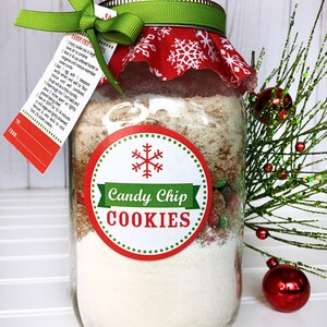 A mason jar filled with cookie mix, labeled "Candy Chip Cookies." The jar is adorned with a red and white snowflake fabric lid cover, a green ribbon, and a tag with instructions. A festive holiday gift.