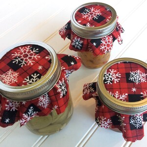 Puede incluir: Tres tarros de vidrio con tapas de tela a cuadros rojos y negros con dise&ntilde;os de copos de nieve blancos. Los tarros est&aacute;n llenos de varios contenidos.