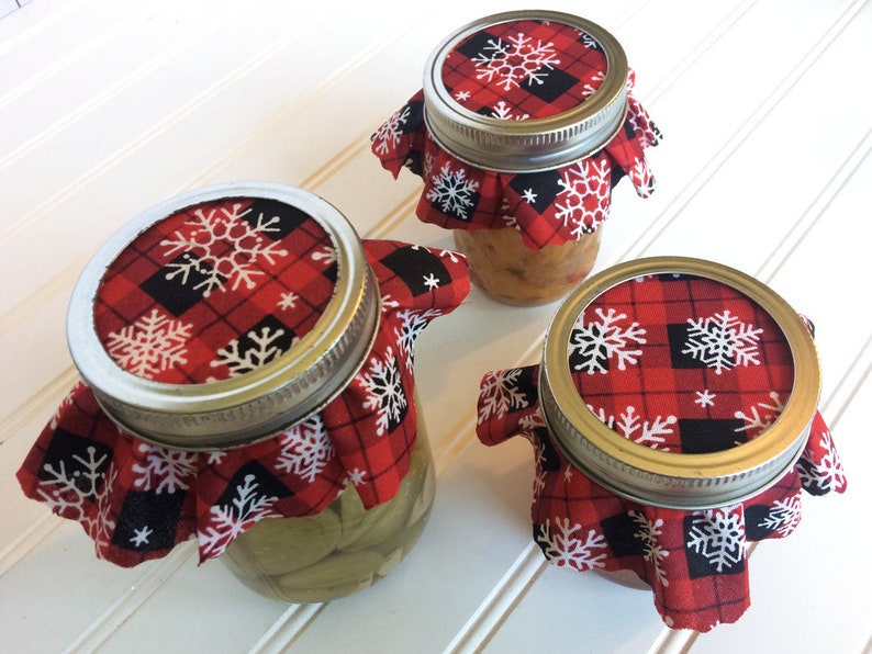 Puede incluir: Tres tarros de vidrio con tapas de tela a cuadros rojos y negros con dise&ntilde;os de copos de nieve blancos. Los tarros est&aacute;n llenos de varios contenidos.