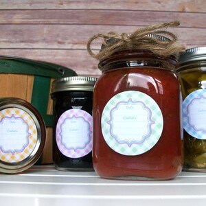 May include: Four glass jars with labels on them. The labels are round and have a gingham pattern. The jars are filled with different types of preserves. The jars are sitting on a white surface.